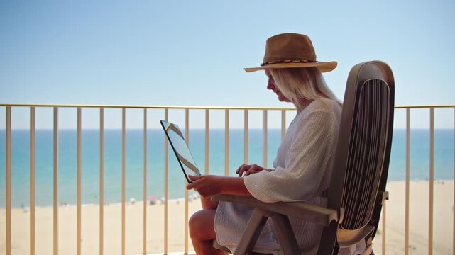 Middle aged woman working on laptop while relaxing on balcony with sea view at sunny beach resort vacation destination