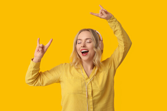 Pretty young woman with modern headphones dancing while listening to music on yellow background