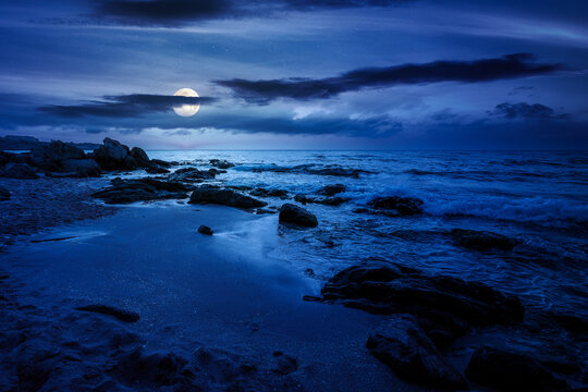 huge stones on a sandy beach at night. wonderful summer vacation season at the black sea. full moon over calm waves washing the sand shore. mysterious nature background with cloudy sky