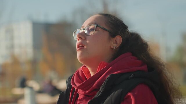 Young woman recording voice on phone, closeup with glasses and hoodie, expressive mouth and focused gaze, vlogger narrating outdoors, singer practicing melody, student leaving voice memo, reflective