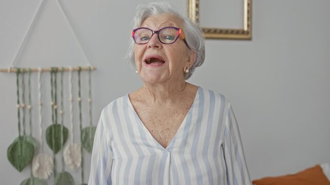 Elderly woman wearing colorful glasses smiles and speaks, standing by macrame wall hanging and cushion in a building living room; warmth joy.