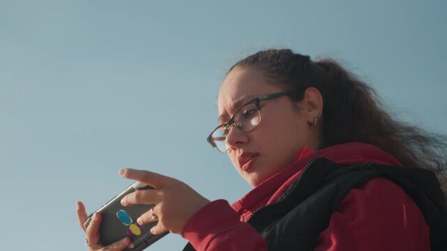 Outdoor side profile woman gaming device, glasses and ponytail, red hoodie, deliberate tapping and swiping gestures, concentrated expressions, clear blue sky and urban lamp posts, close midshot