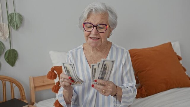 Elderly woman counts dollars, hands fanning bills on a bedroom bed with pillows and macrame wall decor; financial security contentment.