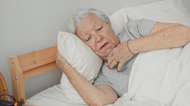Woman clutching chest with hand while reclining on pillow in bed inside a building, red fingernails visible, eyes closed; pain vulnerability.