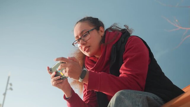 Young woman pointing at smartphone screen while teaching mobile trick on bench, expressive hand gestures, informal recording vibe, urban lamp post and clear sky backdrop, daylight highlights face