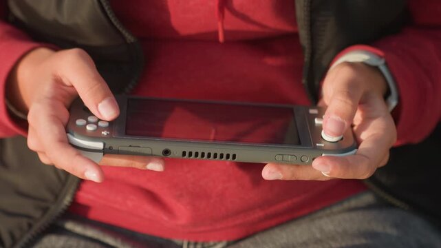 Closeup hands holding handheld console, young player in red hoodie sitting with device on lap, thumbs pressing buttons and joystick, focused concentration and relaxed posture, warm sunlight