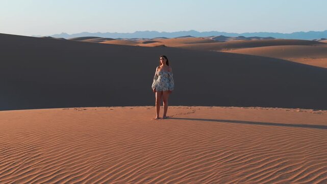 A young women with long hair wearing a summer dress and walking barefoot across vast sand dunes