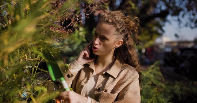 Young woman talking on phone while pruning plants in a sunny garden center, surrounded by greenery and using gardening tools