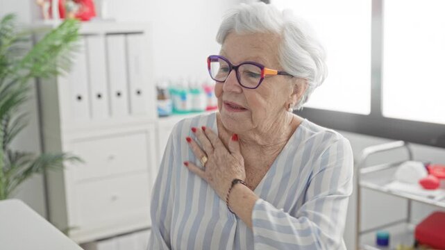 Elderly woman holding chest with hand in clinic building wearing glasses and striped blouse; concern vulnerability.