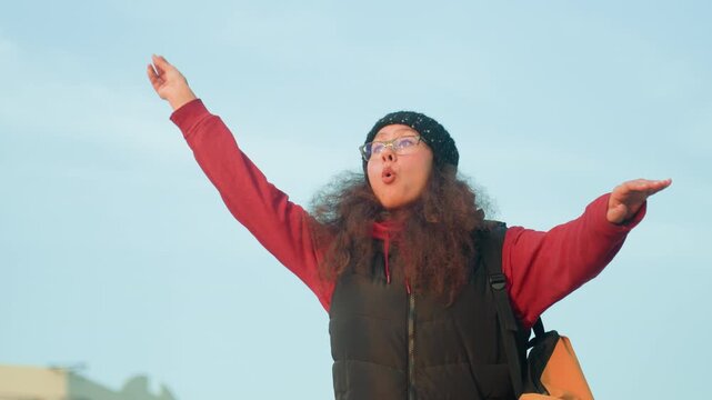 Woman outdoors in winter coat expressive street performer singing and gesturing with beanie and backpack against urban skyline warm sunlight candid smile playful motion conveying freedom and joy