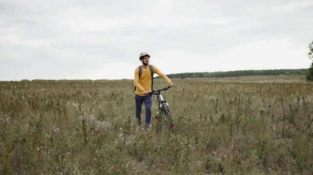 Tired and sad cyclist leading bicycle through wild field among grass. Cyclist lost path, absence of planned route, problem with spatial orientation on spot.