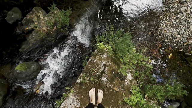 Bare feet rest on a mossy rock above a flowing forest creek with white water moving over dark stones. Pebbles, leafy green plants, shallow water, and natural rock textures create a grounded outdoor se