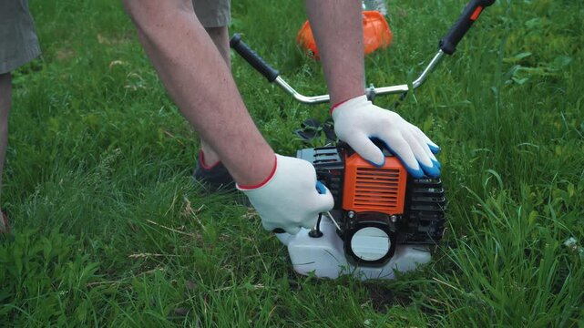 Man in gloves prepares a gas-powered string trimmer for use by checking the engine and adjusting components in a grassy outdoor setting