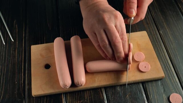 Person slices hot dogs into circular pieces on a wooden cutting board, showcasing a continuous cooking preparation process in a kitchen setting