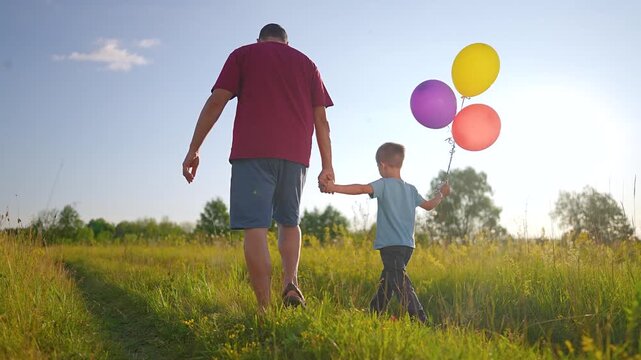 Father and son walk in field holding hands. Boy carries colorful balloons while walking with dad. Parent and child stroll in meadow. Family walks at sunset. Boy holds balloon bundle outdoor.
