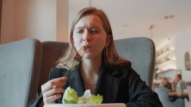 Caucasian woman in cafe grimacing at salad, business attire, fork held hesitantly, pale lettuce on white plate, subtle frown and pursed lips, modern restaurant interior with warm pendant lights,