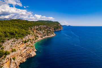 Fototapeta premium Aerial view of rocky coastline with green cliffs and deep blue sea. Dramatic rocky shore covered with lush pine trees next to the crystal clear turquoise and blue waters of Adriatic Sea, Croatia