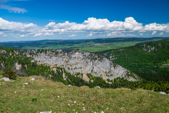 Wide view of limestone cliffs, green forests, and Jura valleys under blue sky and clouds at Creux du Van in spring.