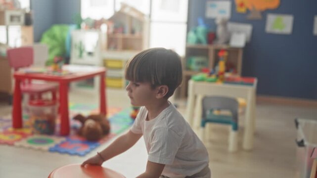 Boy drawing with chalk at wooden easel in a preschool studio among colorful toys, storage bins and small tables; quiet concentration.
