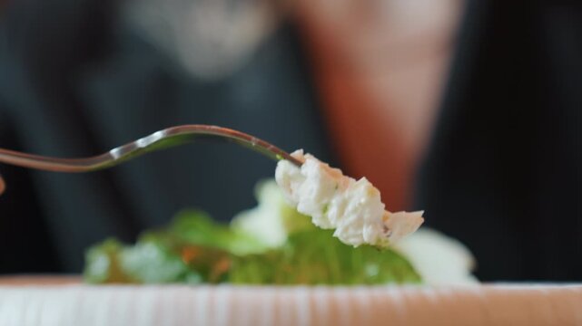 Closeup fork holding salad bite, blurred diner in blazer at upscale restaurant, crisp lettuce and creamy cheese pierced on tines, soft bokeh background, refined tasting atmosphere evoking elegance