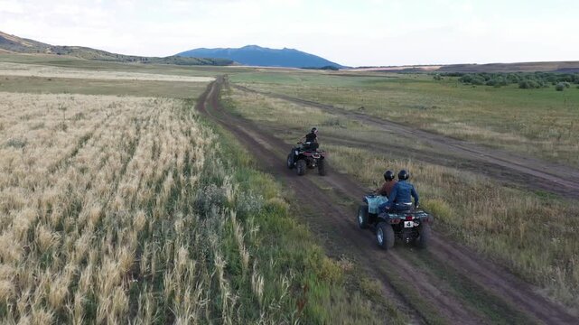 An aerial drone shot of a group of friends riding quad bikes on a dirt path through beautiful green fields, with a scenic lake in the distant background.

