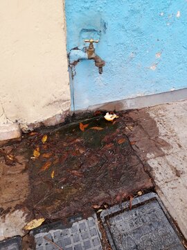 Canilla de agua exterior goteando sobre la vereda urbana junto a una pared azul deteriorada. La p&eacute;rdida  de agua genera barro, humedad y suciedad acumulada con hojas secas alrededor del desague.