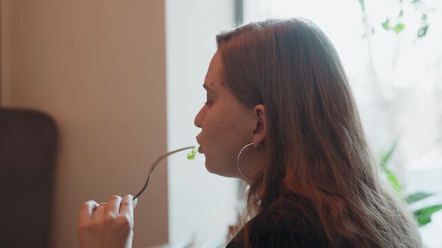 Young caucasian woman sitting by window holding fork gazing thoughtfully during slow morning coffee in cozy bistro soft natural light warm tones hoop earrings long hair quiet mood lifestyle portrait
