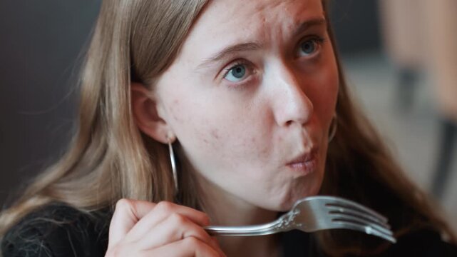 Young woman tasting sour spoon, candid cafe scene showing young adult woman sampling spoonful and making grimace and pensive face, holding metal spoon near mouth, subtle earrings visible, soft natural
