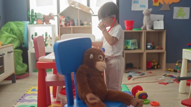 Boy wearing glasses uses red toy stethoscope to check a seated stuffed monkey on a blue chair in building playroom; playful care.
