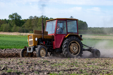 Fototapeta premium Old yellow tractor plowing agricultural field raising dust during spring seasonal farming work with green forest and blue sky in the background countryside