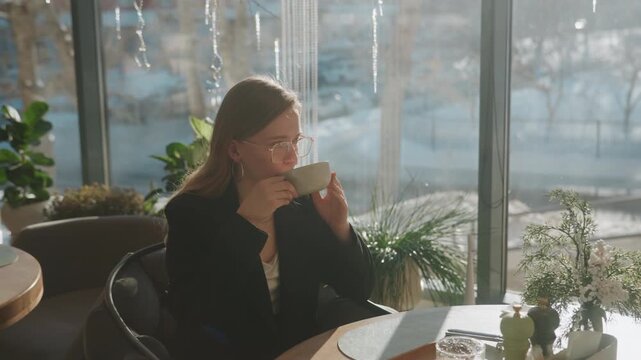 Woman working on laptop in cafe sunlit window overlooking snowy street, notebook and planner on table, blazer and eyeglasses, focused expression, entrepreneur mood, taking coffee breaks, urban