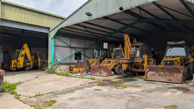 Abandoned industrial site, rusting workshop storage. Rusty excavators, diggers, backhoe loader heavy equipment in depot yard shed. Agricultural worksite vehicle equipment. Disused equipment lying idle