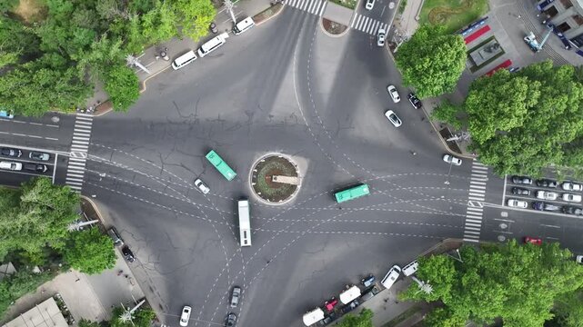An aerial top down view of a busy city intersection with cars moving around a central roundabout surrounded by lush green trees on a bright sunny summer day.

