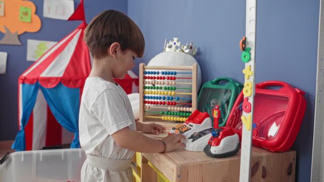 Boy points finger at a red toy cash register and presses buttons with his bare hand in a playroom building; curiosity learning.