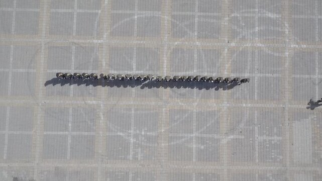 Top down aerial view of military soldiers marching in a synchronized formation on a paved city square during a parade, creating intricate geometric patterns and casting long shadows.

