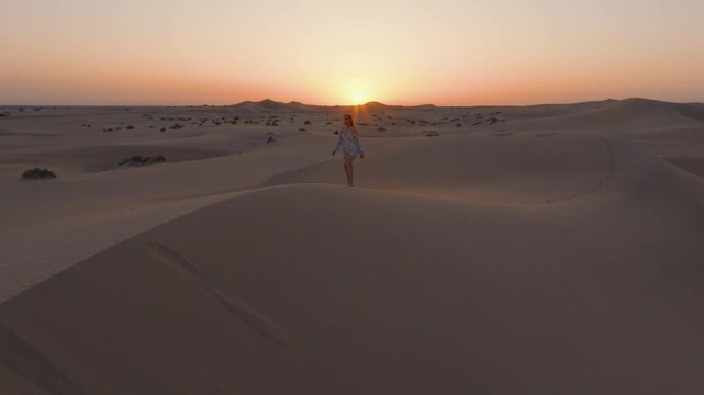 A young women with long hair wearing a summer dress and walking barefoot across vast sand dunes