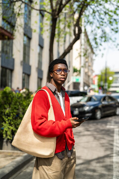 Focused african american man student holding smartphone in hand standing on street waiting for taxi, thoughtful fashionable black male outdoors ordering car using phone app, guy waiting for transport.