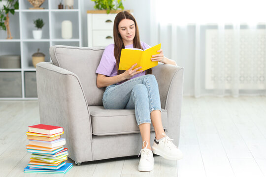 Young woman reading book on armchair near stack of colorful ones at home. Space for text