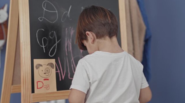 Boy drawing on a chalkboard at a wooden easel in a playroom setting, back to camera in a white tshirt, holding chalk and sketching letters and a dog card; curiosity learning.
