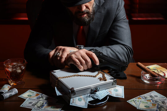 Mafia. Man with gun, case of dollar banknotes and golden chain at wooden table in dark room