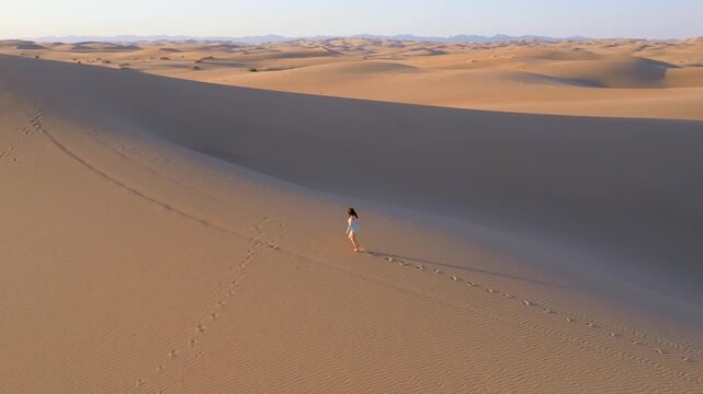 A young women with long hair wearing a summer dress and walking barefoot across vast sand dunes