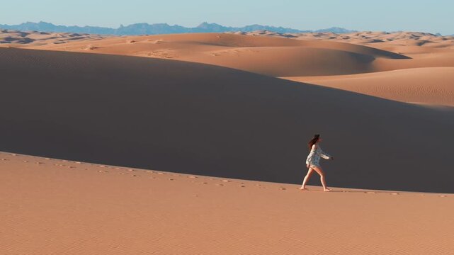 A young women with long hair wearing a summer dress and walking barefoot across vast sand dunes