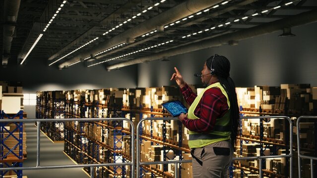 African American female staff using headset to handle package tracking, cargo status and customer complaints on a call, Woman transmitting parcel tracking info for e-commerce services.