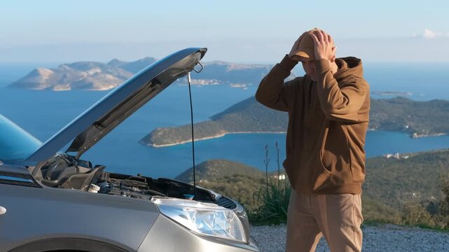 Stressed man standing near broken down car. Frustrated man looking at his broken car engine with the hood open, clutching his head in despair during a scenic road trip with a beautiful sea
