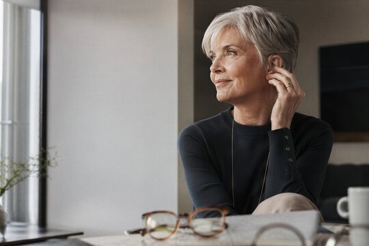 Senior woman with short grey hair adjusting a discreet hearing aid. Happy elderly lady using modern medical device for hearing loss, enjoying communication and life. Clear wellness concept
