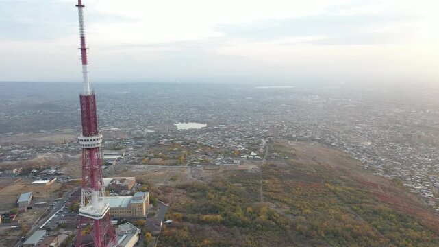 An aerial drone view of the Yerevan TV Tower standing tall over the misty Armenian capital during autumn with colorful forest and scenic city panorama in the background.

