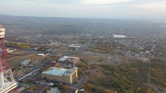 An aerial drone view of the Yerevan TV Tower standing tall over the misty Armenian capital during autumn with colorful forest and scenic city panorama in the background.

