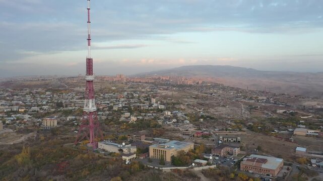 An aerial drone view of the Yerevan TV Tower standing tall over the misty Armenian capital during autumn with colorful forest and scenic city panorama in the background.

