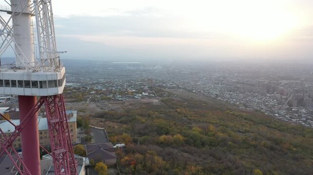 An aerial drone view of the Yerevan TV Tower standing tall over the misty Armenian capital during autumn with colorful forest and scenic city panorama in the background.

