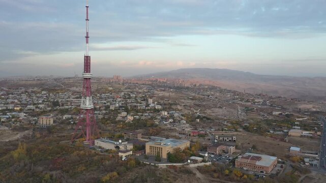 An aerial drone view of the Yerevan TV Tower standing tall over the misty Armenian capital during autumn with colorful forest and scenic city panorama in the background.

